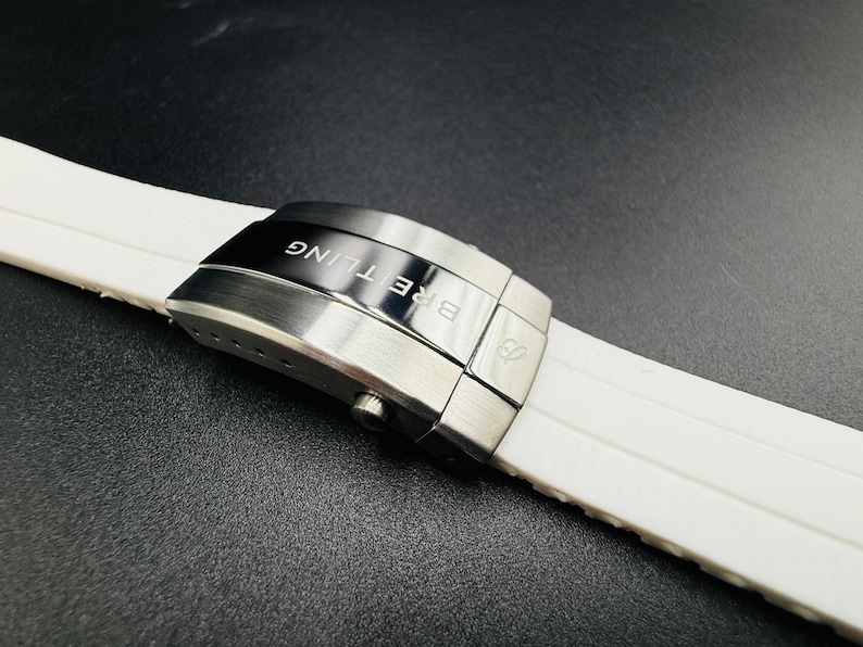 Puede incluir: Primer plano de una correa de reloj blanca con un cierre plateado. El cierre est&aacute; grabado con la palabra "BREITLING" en negro. La correa est&aacute; hecha de un material suave y flexible.