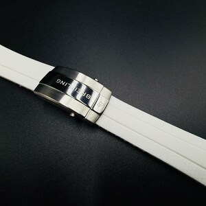 Puede incluir: Correa de reloj blanca con cierre de acero inoxidable. El cierre est&aacute; grabado con la palabra "Breitling". La correa est&aacute; hecha de un material suave y flexible.