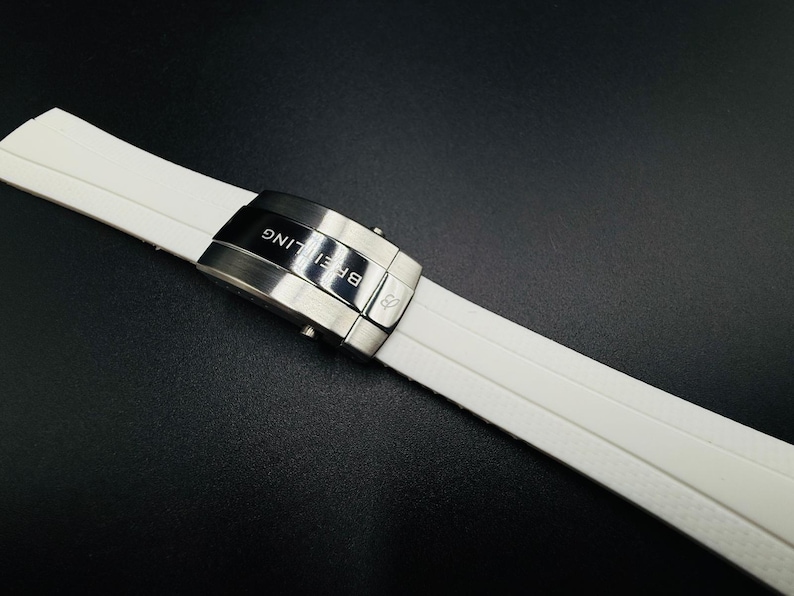 Puede incluir: Correa de reloj blanca con cierre de acero inoxidable. El cierre est&aacute; grabado con la palabra "Breitling". La correa est&aacute; hecha de un material suave y flexible.