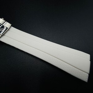 Puede incluir: Primer plano de una correa de reloj blanca con un cierre plateado. La correa est&aacute; hecha de un material liso y flexible con una parte inferior texturizada. El cierre tiene grabado el nombre de la marca "BREITLING".