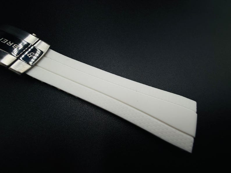 Puede incluir: Primer plano de una correa de reloj blanca con un cierre plateado. La correa est&aacute; hecha de un material liso y flexible con una parte inferior texturizada. El cierre tiene grabado el nombre de la marca "BREITLING".