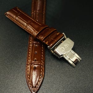May include: A brown leather watch strap with a crocodile pattern. The strap features a stainless steel clasp with the letters "IWC" engraved. The watch strap is new and unworn.