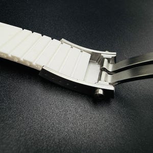 Puede incluir: Primer plano de una correa de reloj blanca con un cierre de acero inoxidable. La correa tiene un dise&ntilde;o texturizado y acanalado. El cierre est&aacute; abierto, revelando su mecanismo interno. El fondo es una superficie oscura y texturizada.