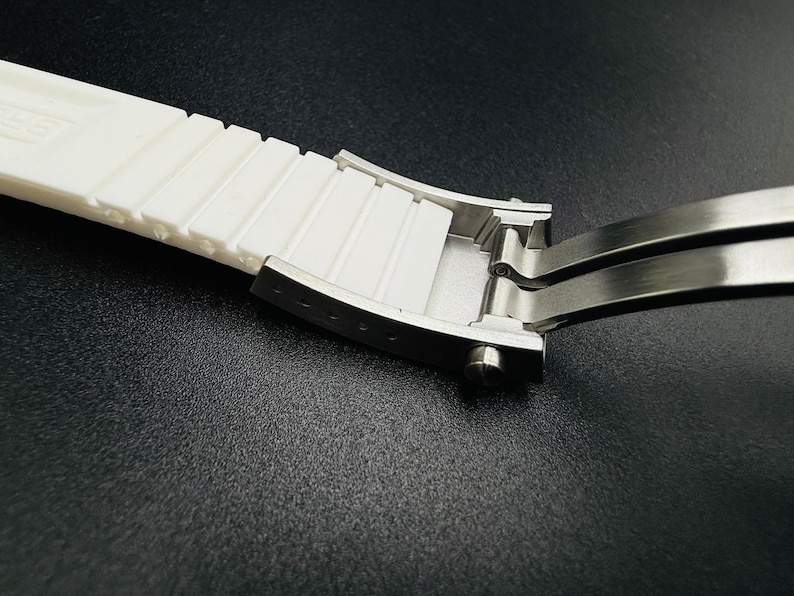 Puede incluir: Primer plano de una correa de reloj blanca con un cierre de acero inoxidable. La correa tiene un dise&ntilde;o texturizado y acanalado. El cierre est&aacute; abierto, revelando su mecanismo interno. El fondo es una superficie oscura y texturizada.