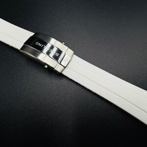 Puede incluir: Correa de reloj blanca con cierre plateado. La correa est&aacute; hecha de un material texturizado y presenta la marca "Breitling". El cierre es plateado y tiene el nombre de la marca grabado. La correa est&aacute; sobre una superficie negra.