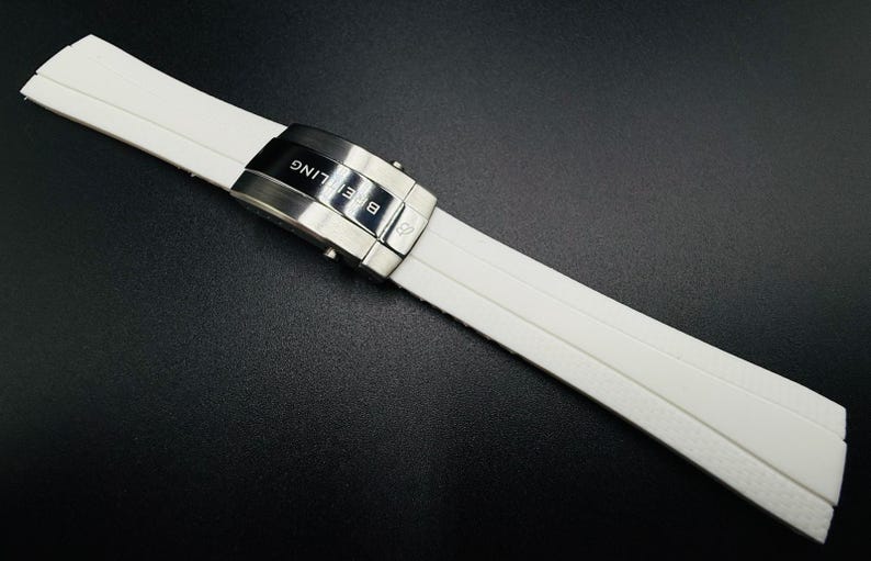 Puede incluir: Correa de reloj blanca con cierre plateado. La correa est&aacute; hecha de un material texturizado y presenta la marca "Breitling". El cierre es plateado y tiene el nombre de la marca grabado. La correa est&aacute; sobre una superficie negra.