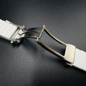 Puede incluir: Primer plano de una correa de reloj blanca con un cierre de acero inoxidable. La correa es de un material texturizado y acanalado. El cierre est&aacute; abierto, revelando el mecanismo. El fondo es una superficie oscura y texturizada.