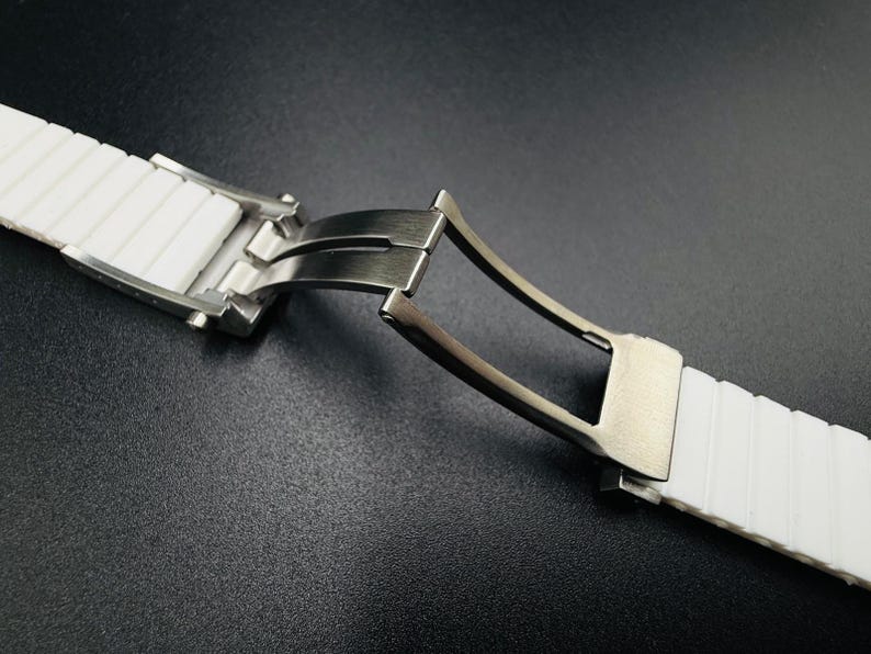 Puede incluir: Primer plano de una correa de reloj blanca con un cierre de acero inoxidable. La correa es de un material texturizado y acanalado. El cierre est&aacute; abierto, revelando el mecanismo. El fondo es una superficie oscura y texturizada.