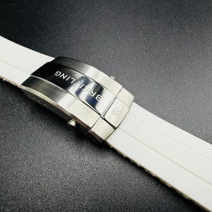 Puede incluir: Primer plano de una correa de reloj blanca con un cierre de acero inoxidable. El cierre est&aacute; grabado con el nombre de la marca "Breitling" en letras blancas. La correa del reloj est&aacute; hecha de un material texturizado y acanalado.