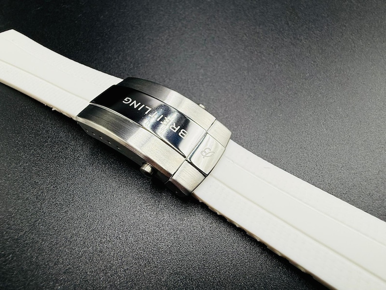 Puede incluir: Primer plano de una correa de reloj blanca con un cierre de acero inoxidable. El cierre est&aacute; grabado con el nombre de la marca "Breitling" en letras blancas. La correa del reloj est&aacute; hecha de un material texturizado y acanalado.