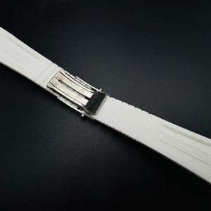 Puede incluir: Una correa de reloj blanca con un cierre plateado. La correa est&aacute; hecha de un material texturizado y acanalado y presenta el nombre de la marca "BREITLING" en relieve. La correa est&aacute; dise&ntilde;ada para sujetarse a un reloj.