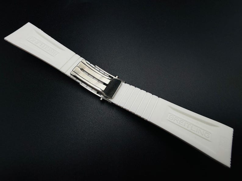 Puede incluir: Una correa de reloj blanca con un cierre plateado. La correa est&aacute; hecha de un material texturizado y acanalado y presenta el nombre de la marca "BREITLING" en relieve. La correa est&aacute; dise&ntilde;ada para sujetarse a un reloj.