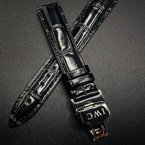 May include: Black leather watch straps with a crocodile pattern. The straps are glossy and feature a silver clasp with the letters "IWC" engraved. The watch straps are designed to be durable and stylish.