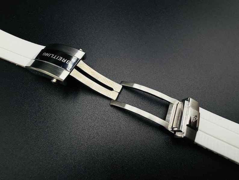 Puede incluir: Primer plano de una correa de reloj blanca con un cierre plateado. El cierre est&aacute; grabado con la marca "Breitling" en negro. La correa est&aacute; hecha de un material suave y flexible.