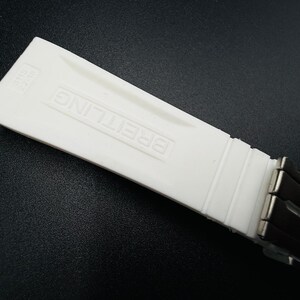 Puede incluir: Un componente de correa de reloj blanco con el nombre de la marca "BREITLING" en relieve. La correa est&aacute; hecha de un material liso y mate y est&aacute; conectada a una secci&oacute;n de correa negra con cierres met&aacute;licos. El fondo es una superficie oscura.