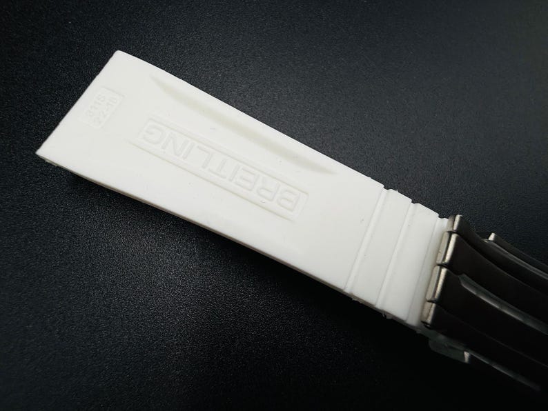 Puede incluir: Un componente de correa de reloj blanco con el nombre de la marca "BREITLING" en relieve. La correa est&aacute; hecha de un material liso y mate y est&aacute; conectada a una secci&oacute;n de correa negra con cierres met&aacute;licos. El fondo es una superficie oscura.