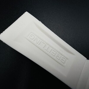 Puede incluir: Primer plano de una correa de reloj blanca con la palabra "BREITLING" en relieve. La correa est&aacute; hecha de un material flexible y tiene una superficie texturizada. El fondo es negro.