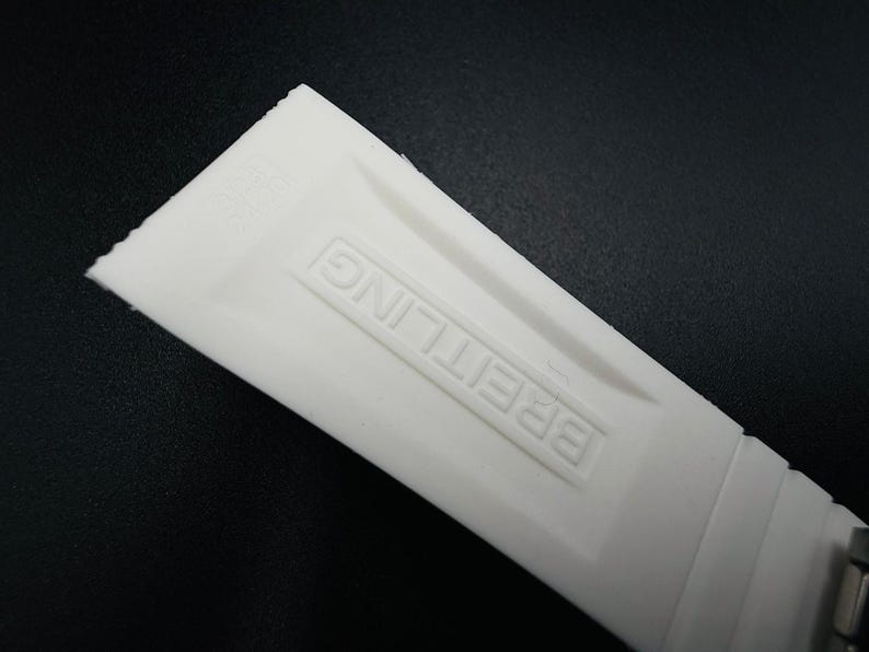Puede incluir: Primer plano de una correa de reloj blanca con la palabra "BREITLING" en relieve. La correa est&aacute; hecha de un material flexible y tiene una superficie texturizada. El fondo es negro.