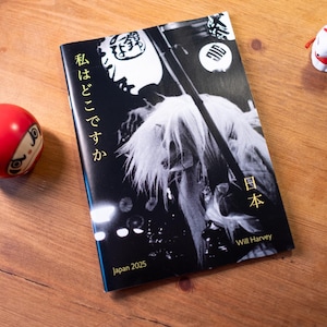May include: A book titled "Japan 2025" by Will Harvey, featuring a black and white image of a person with long hair. A red Daruma doll and a white fox figurine are also present on a wooden surface.