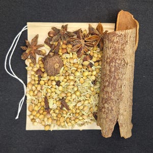 May include: A spice blend featuring star anise, coriander seeds, cloves, and a cinnamon stick. The blend is presented on a small, square, light brown paper packet with a white string attached. The spices are a mix of brown, tan, and beige.