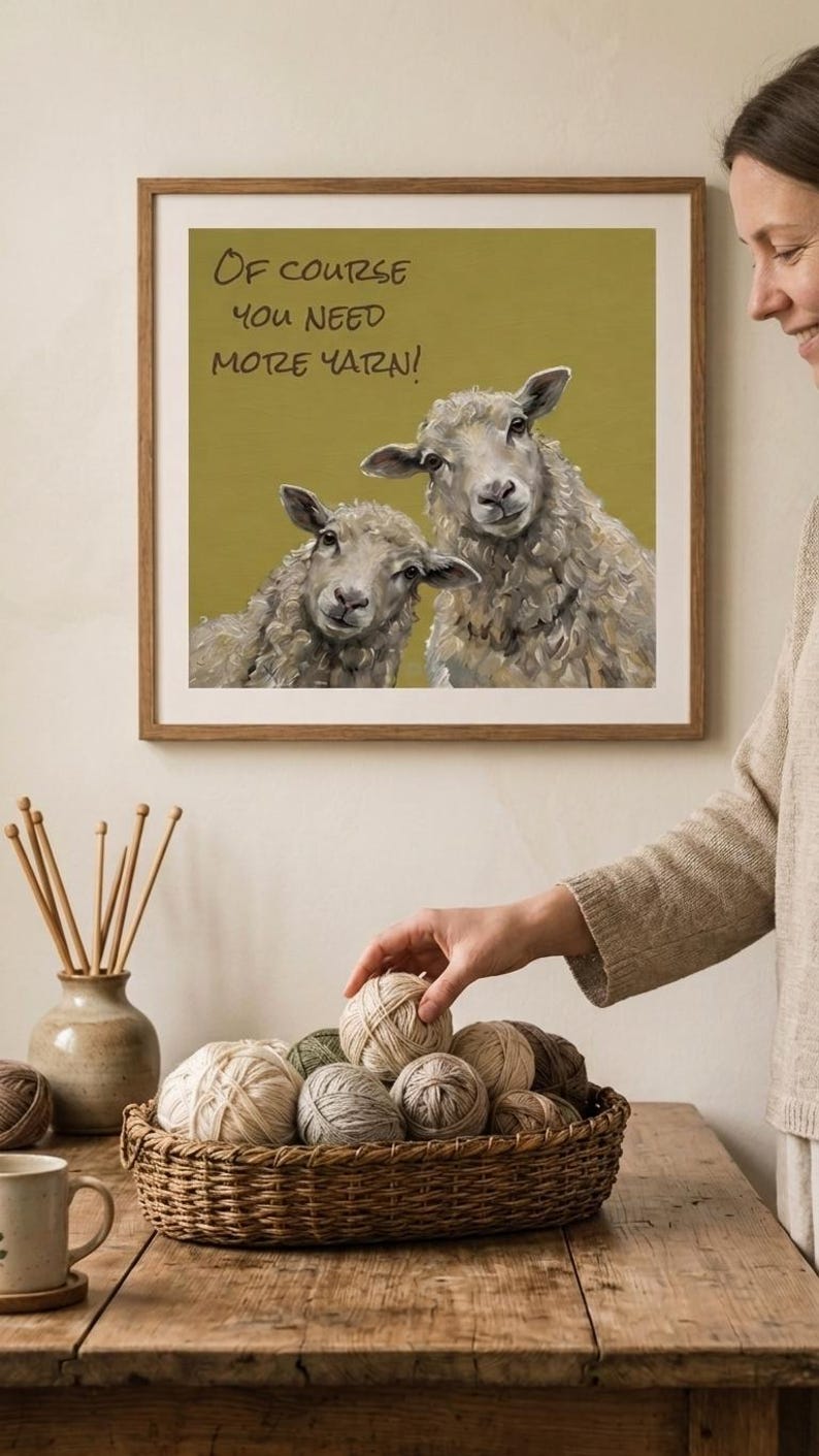 May include: A framed wall print featuring an oil-style painting of two sheep against an olive green background with the text "OF COURSE YOU NEED MORE YARN!" in black. The print is displayed in a simple wooden frame on a light-colored wall.