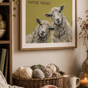 May include: A square framed wall art print featuring an olive green background with two sheep and the text "Of course you need more yarn!" in black lettering. The artwork is displayed in a light wood frame, showcasing a detailed painting of sheep.