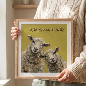 May include: A square framed print featuring an oil painting of two sheep against a mustard yellow background. The text "ARE YOU KNITTING?" is printed in black at the top. The sheep have textured, off-white wool and are looking forward.