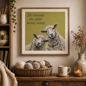 May include: A square framed wall art print featuring an olive green background with two sheep and the text "Of course you need more yarn!" in black lettering. The sheep are rendered in shades of gray, white, and brown with visible brushstrokes.