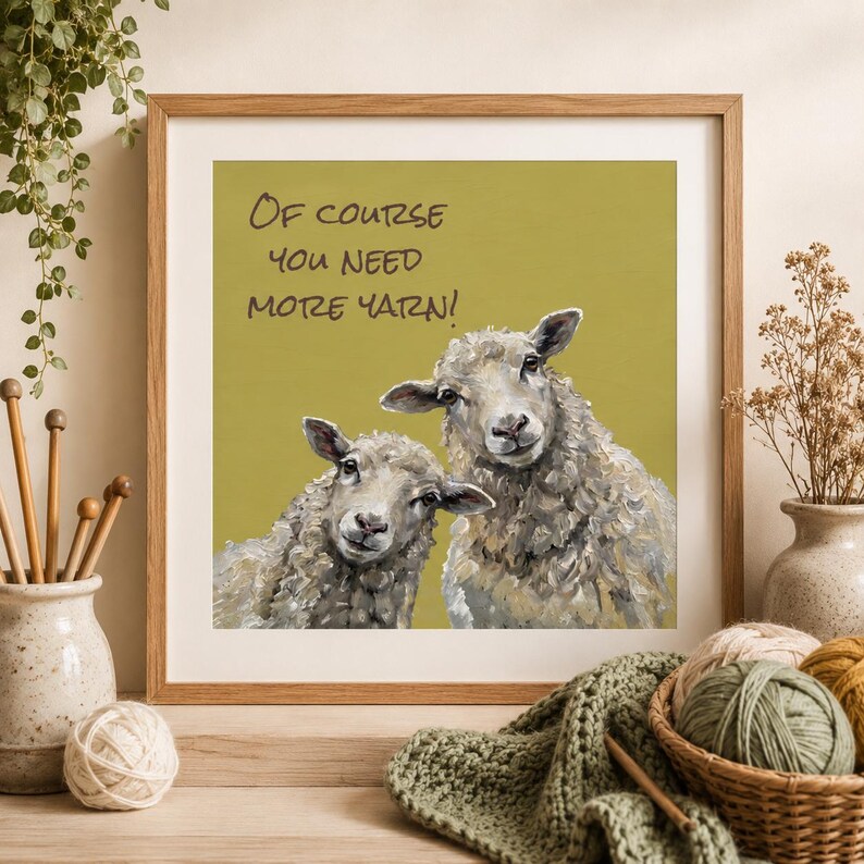 May include: A square framed wall art print featuring an olive green background with two textured, painted sheep. The text "OF COURSE YOU NEED MORE YARN!" is printed in black above the sheep. The artwork is displayed in a light wood frame.