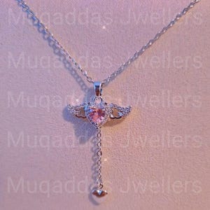 Op de afbeelding: Een zilveren ketting met een hanger met een roze hartvormige edelsteen, omringd door kleine heldere stenen en vleugels. Een ketting loopt naar beneden en eindigt met een klein hartje. De achtergrond is zachtroze.