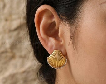 May include: A gold-colored seashell-shaped earring with a row of small, clear stones along the bottom edge. The earring is worn on an ear, and the background is blurred.