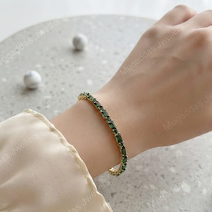 Könnte beinhalten: Ein goldfarbenes Tennisarmband mit einer durchgehenden Reihe rechteckiger, smaragdgrüner Edelsteine in einer Metallfassung. Das Armband wird am Handgelenk getragen und präsentiert die leuchtend grünen Steine sowie die goldene Metallfassung.