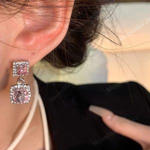 May include: A silver-toned drop earring featuring two square-shaped components. The top component is a small square with a pink stone, and the bottom component is a larger square with a round pink stone, both bordered by small clear crystals.