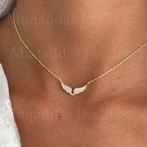 May include: A gold necklace with a pendant in the shape of angel wings, adorned with small crystals. The necklace is shown on a person's skin, showcasing its elegant design. This piece of jewellery is a stylish accessory.