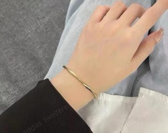 May include: A thin, gold-toned metal bangle bracelet featuring a continuous, gentle wave or twisted design. The bracelet has a smooth, polished surface and a circular shape that rests around the wrist, showcasing a simple and sleek aesthetic.