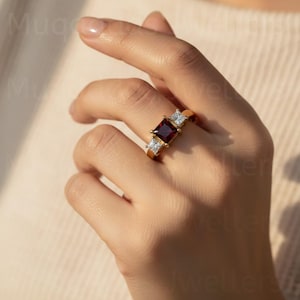 May include: A gold ring with a central, square, deep red gemstone, and two smaller, square, clear gemstones. The ring is displayed on a hand, highlighting the jewellery's design and colour contrast.