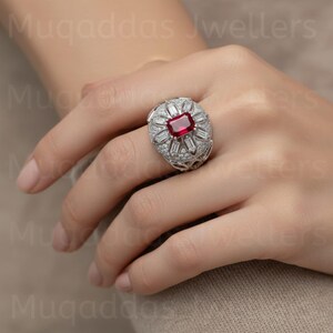 May include: A silver ring with a rectangular, deep red gemstone at the center, surrounded by smaller, clear stones. The ring has a detailed, ornate design. The image is of a hand wearing the ring.