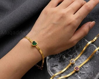 May include: A gold-toned flat snake chain bracelet featuring a single green heart-shaped stone. A second matching gold-toned chain bracelet is visible, adorned with two small heart-shaped stones in clear and pink hues, set along the chain.