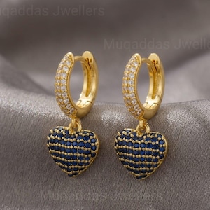May include: A pair of gold-colored hoop earrings. The top portion of each earring is encrusted with small, clear stones. Each earring has a heart-shaped pendant covered in small, dark blue stones. The earrings are displayed on a gray surface.
