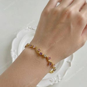 May include: A gold-toned bracelet featuring a delicate, wavy band design. The bracelet is adorned with a series of small, round-cut gemstones in varying shades of pink, purple, and clear white, arranged along the undulating metal structure.