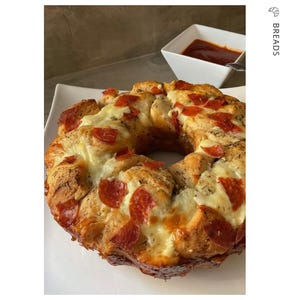 May include: A baked pizza ring with pepperoni and melted cheese on a white plate. A small white bowl of red sauce is in the background. The word "BREADS" is printed vertically on the right side of the image.
