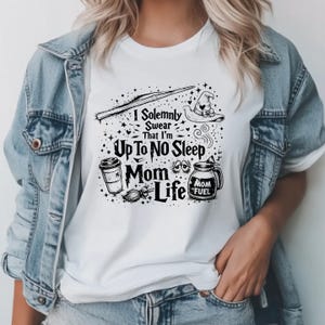 May include: White t-shirt with a black graphic design featuring a wand, witch hat, coffee cup, and the text "I Solemnly Swear That I'm Up To No Sleep Mom Life". The shirt is paired with a denim jacket.