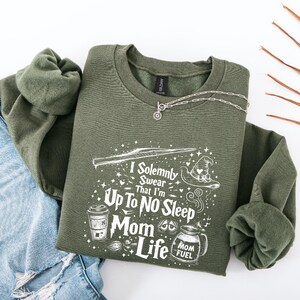 May include: An olive green sweatshirt with the text "I Solemnly Swear That I'm Up To No Sleep Mom Life" in white. The design includes a wand, a witch's hat, a coffee cup, and a jar labeled "Mom Fuel". A silver necklace is draped across the sweatshirt.