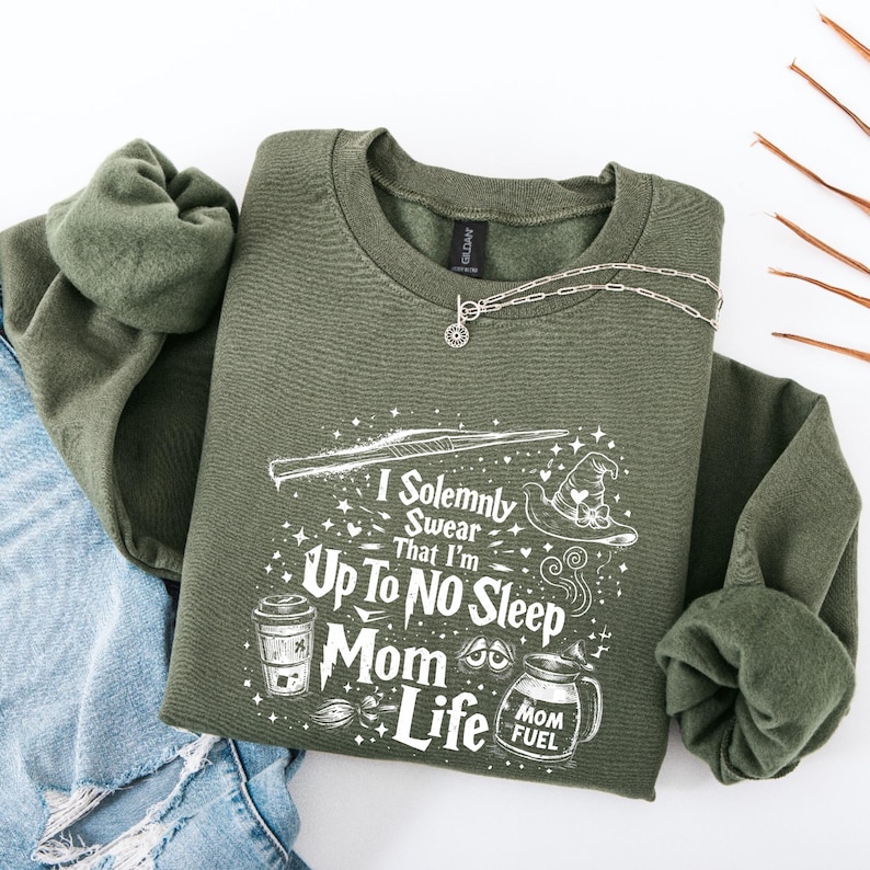 May include: An olive green sweatshirt with the text "I Solemnly Swear That I'm Up To No Sleep Mom Life" in white. The design includes a wand, a witch's hat, a coffee cup, and a jar labeled "Mom Fuel". A silver necklace is draped across the sweatshirt.