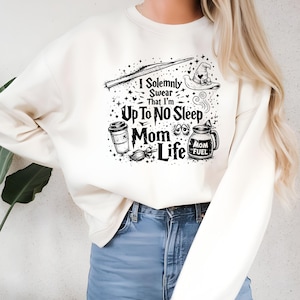 May include: Cream-colored sweatshirt with a black graphic design. The design includes a wand, witch hat, coffee cup, broom, and the text "I Solemnly Swear That I'm Up To No Sleep Mom Life." The sweatshirt is paired with blue jeans.