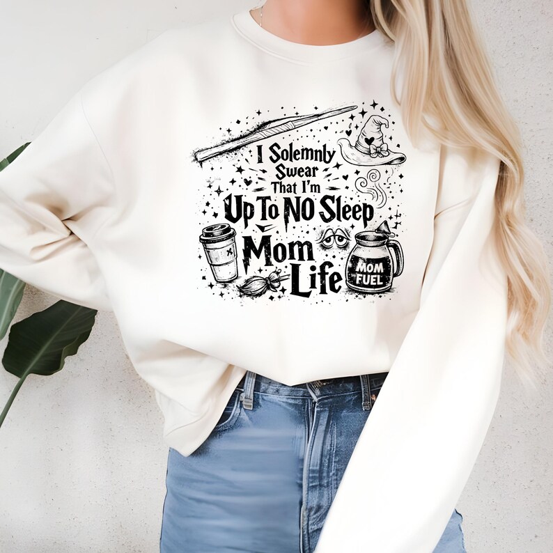 May include: Cream-colored sweatshirt with a black graphic design. The design includes a wand, witch hat, coffee cup, broom, and the text "I Solemnly Swear That I'm Up To No Sleep Mom Life." The sweatshirt is paired with blue jeans.