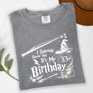May include: Gray t-shirt with a white graphic design featuring a wand, stars, a wizard hat, and the text "I Solemnly Swear that It's My 33rd Birthday". The shirt has a crew neck and a tag that says "Comfort Colors".