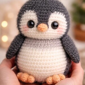 Puede incluir: Un pingüino de ganchillo hecho a mano con cabeza y alas grises, vientre blanco y pico y pies naranjas. El pingüino tiene grandes ojos negros y mejillas rosadas. Un peluche suave y tierno.