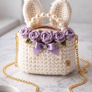 May include: A cream-colored crochet handbag with bunny ears and a pearl handle. The bag features a row of purple crochet roses, a lavender bow with a rhinestone, and a gold chain strap. A button closure is visible.