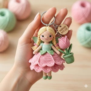 Puede incluir: Un llavero de hada de ganchillo hecho a mano con un vestido rosa y trenzas rubias. El hada sostiene un tulipán rosa en una maceta verde. Una pequeña etiqueta de madera dice "HANDMADE WITH LOVE".
